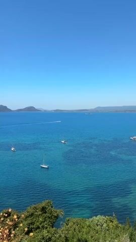 The video shows a wide-angle shot of a bay with a clear blue sky overhead. The water is turquoise and relatively calm, with visible underwater patterns. Several sailboats and a powerboat are scattered across the water, appearing stationary. In the distance, land is visible, including a small island and a coastline with low hills. The foreground features green vegetation, suggesting a coastal vantage point. The camera is steady, providing a static view of the scene.