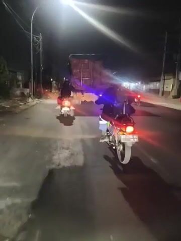 The video shows a nighttime view of a road with several motorcycles and a large truck. The motorcycles are moving in the same direction as the truck. The truck appears to be stopped or moving very slowly, as the motorcycles are able to pass it. The road is paved and well-lit by streetlights and the headlights of the vehicles. There are buildings and trees visible on the sides of the road. The overall impression is of a group of motorcyclists navigating through traffic at night.