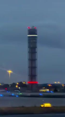 The video shows a tall, slender tower at dusk, with red lights at the top and around the middle. The tower appears to be made of concrete and metal, and is located near an airport runway with blue and green lights visible. In the background, there are scattered city lights and a streetlight. The sky is cloudy and dark.