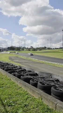 A white race car with red and orange accents speeds along a paved racetrack. The car is positioned on the right side of the track as it moves away from the camera. In the foreground, a barrier made of stacked tires secured with chains runs along the edge of the track, separated by a concrete curb. Beyond the track, a grassy field is visible, with trees and a building in the distance. The sky is a mix of blue with scattered white clouds. Another white car is further down the track in the distance.