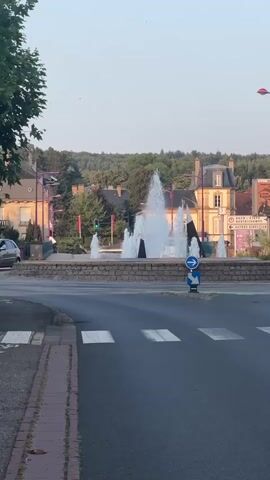 The video shows a fountain in a town square. The fountain is spraying water into the air, and there are trees and buildings in the background. A crosswalk is visible in the foreground, and a street sign with a blue arrow indicating a roundabout is present. A few people are seen walking around the fountain and in the distance. The sky is clear and the lighting suggests it is daytime.