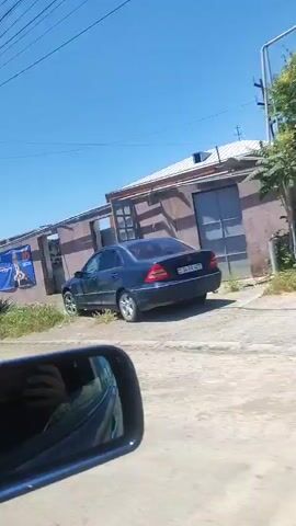 A video shows a moving vehicle's perspective from the passenger side, looking out at a roadside environment on a clear, sunny day. A blue side mirror in the bottom left reflects a hand holding a phone, indicating the recording device. The vehicle travels along a paved road. On the right, there are various structures including a building with a large blue banner displaying a man in a suit forming a heart with his hands and text in an unknown language along with the number "16". Further along the road, stone walls, green trees, bushes, a wooden utility pole with wires, and a reddish-brown metal gate are visible.