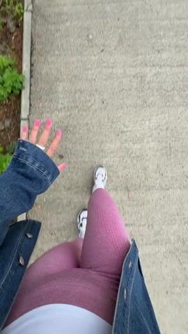 This is a point-of-view video of a person walking on a paved path. The person is wearing a denim jacket, pink leggings, white sneakers, and a white top. Their fingernails are painted pink. The path is bordered by grass and trees. In the background, there are buildings and a cloudy sky. The person waves at the camera and makes a peace sign.
