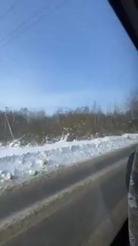 The video shows a roadside view from a moving vehicle on a sunny winter day. The road is mostly clear of snow but wet. Heavy snow covers the ground next to the road and extends into the bare-branched trees and bushes. The sky is clear and bright blue.