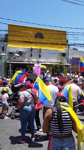 Here's a concise description of the video: 

The video shows a daytime street scene where a crowd of people are walking and celebrating. Many are carrying and waving Venezuelan flags. Most of the people appear to be dressed casually. The buildings visible on the sides of the street are mostly low-rise, with a few taller buildings visible in the distance. The weather is sunny and the sky is clear. There are some utility poles and wires overhead.  The general atmosphere is festive and celebratory.