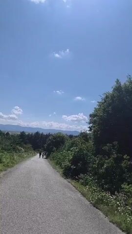 The video opens with a wide shot of a path leading towards a group of people in the distance, surrounded by lush green trees under a clear blue sky. The camera then pans across the mountainous landscape, revealing more trees, distant hills, and a bright blue sky dotted with white clouds.