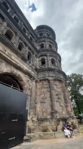 The video shows a low-angle, upward view of a large, ancient stone structure, possibly a gate or tower, with multiple levels and arched openings. The stonework is weathered and appears to be very old. In the foreground, partially obscuring the lower part of the structure, is a large, black, rectangular panel with horizontal seams and visible hardware, possibly a temporary construction barrier or a protective cover. To the right of this panel, two people are seated close together on a low stone ledge, facing away from the camera. The person on the left is wearing a blue shirt and white shorts, and the person on the right is wearing a dark shirt and jeans. Behind them, a tree with green foliage is visible. The sky above is cloudy with patches of blue and a wispy white cloud formation. The camera pans slightly to the left and then to the right, revealing more of the structure's facade. As the camera moves, more people come into view walking through the arched entryway of the structure, suggesting it is a public space or monument. The lighting is natural, indicating it is daytime. The overall impression is of a historical site with some ongoing work or preservation efforts.