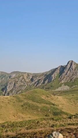 The video pans across a mountainous landscape. The foreground is covered with dry grass and scattered rocks. In the middle ground, rolling hills are covered in a mix of dry and green grass, with some patches of shrubbery. The background features rugged mountain ranges with rocky outcrops and green vegetation. The sky is clear and blue.