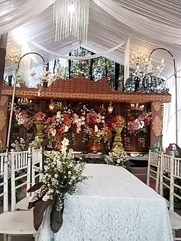 This is a video of an outdoor event space, likely a wedding or reception, with a tent overhead. The area is decorated with flowers and elaborate traditional structures. In the foreground, a table is draped with a white lace tablecloth. To the left, a large arrangement of white flowers with brown accents is visible. Behind the table, there is a decorated wooden structure with cascading red and pink flowers. To the right of this structure, chairs with white cushions are arranged. Further in the background, a rustic, thatched-roof structure with banana trees and other foliage is prominent. Some people are visible in the distance, standing near doorways and within the structures. The overall impression is a festive and culturally inspired setting.
