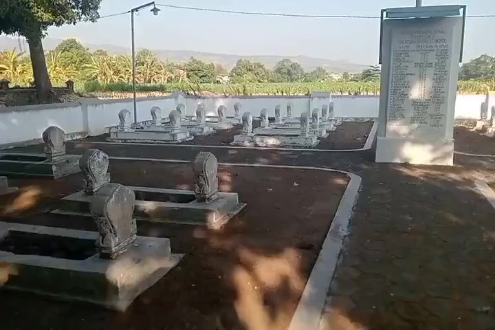 The video shows a cemetery with rows of white gravestones and a central monument with names inscribed on it. The ground is covered with brown soil and some paved pathways. In the background, there are trees, buildings with distinctive roofs, and a fence. The sky is clear and blue, suggesting it is daytime. The scene appears tranquil and well-maintained.