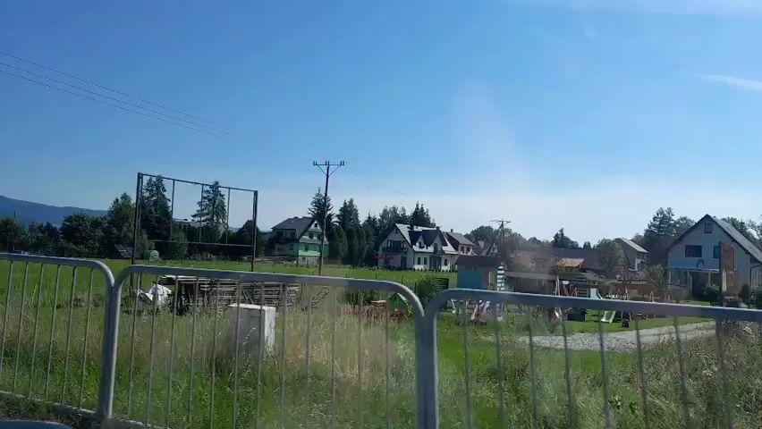 The video captures a daytime view of a rural or suburban landscape from a moving vehicle. In the foreground, a metal fence runs horizontally across the bottom of the frame, with tall grass and vegetation visible behind it. Beyond the fence, a green field stretches out, dotted with some piles of wood and small structures. In the mid-ground, there's a sports court, possibly a tennis court or volleyball court, with a net and poles. Several houses with pitched roofs are visible further back, nestled among trees. The sky is clear and blue. The video appears to be a continuous shot, with no apparent editing or special effects.