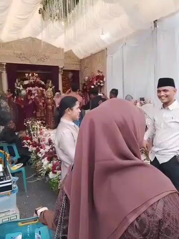 The video shows a wedding ceremony taking place inside a decorated hall. A bride and groom are seated on a raised platform at the far end of the hall, surrounded by floral decorations. In the foreground, several guests are milling about, some engaged in conversation and others observing the ceremony. A woman in a brown hijab is standing with her back to the camera, facing the wedding party. A man in a striped shirt and a black cap is smiling and looking towards the wedding party. There is a large speaker and some audio equipment on the left side of the frame. The hall is decorated with white drapes and elaborate floral arrangements. The overall atmosphere appears to be festive and celebratory.