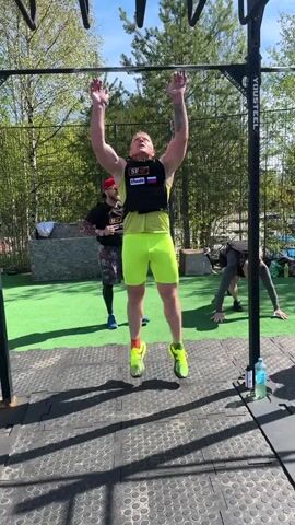 A man in a black weight vest, bright yellow shorts, and neon green shoes, performs assisted pull-ups outdoors. He hangs from a black steel pull-up bar affixed to a metal structure with the word "YOUSTEEL" on it. Another man stands behind him, providing assistance by holding his ankles during the pull-up. A third person, out of focus, performs push-ups on the artificial turf in the background. The location appears to be an outdoor gym or training area, with a green artificial turf and black rubber mats around the pull-up structure. Trees are visible in the background, and the weather appears to be sunny. A bottle of water stands on the ground near the pull-up station.