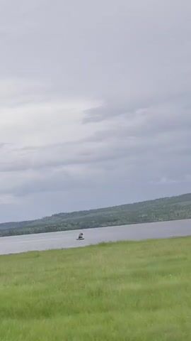 A distant view shows a wide body of water with a treed shoreline under a cloudy sky. In the foreground, green grass covers a slight incline. Several small boats are visible on the water.