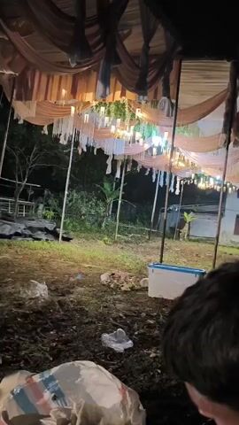 The video opens with a low-angle shot looking up at a tent decorated with fabric drapes and lights. The ground beneath the tent is uneven and appears to be dirt with some scattered debris, including plastic bags. In the distance, a building with lights on is visible, and trees and foliage can be seen behind it.

As the video progresses, the camera pans to the right, revealing more of the tent structure and the surrounding area. There is a white cooler on the right side of the frame. The ground continues to be littered with debris.

The camera then shifts focus to people. In the foreground, a person's back is visible, wearing a white and black shirt. They are bent over, possibly interacting with something on the ground. To their right, a person is seated on a blue chair, looking at a phone. The background shows a building with windows and doors, illuminated by artificial lights.

The video continues to show the scene from different angles, focusing on the people present and the makeshift tent setup. There are more blue chairs visible, and the ground remains somewhat unkempt. Motorcycles are parked in the background. The overall impression is of an outdoor gathering or event, possibly in a semi-rural or informal setting, taking place at night.