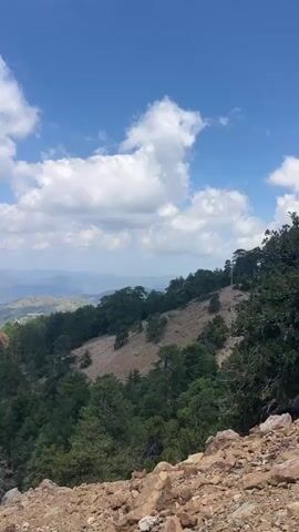 This is a vertical, handheld video of a mountainous landscape. The sky is blue with scattered white clouds. In the foreground, there are rocks and trees, with a steep, rocky slope to the right. The middle ground is filled with a dense forest of evergreen trees. In the background, there are rolling hills and mountains extending into the distance, with a hazy, bluish hue. A small settlement or village can be seen nestled in the valley. The video pans slightly to the right, revealing more of the mountain range. The overall impression is of a natural, serene environment.