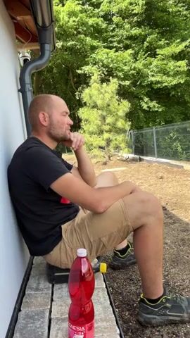 A man with a shaved head and a beard is seated outdoors, leaning against a white wall in Nový Jáchymov, Czechia. He is wearing a black t-shirt, khaki shorts, and black and yellow sneakers. His chin rests on his hand as he looks thoughtfully to his right (the viewer's left).

In front of him, on the paved ground, is a red plastic water bottle with "MAGNESIA" visible on its label. A gray downspout runs vertically along the white wall next to him. The background features lush green trees and foliage, with a fence visible further back, indicating a garden or natural area. The scene is brightly lit, suggesting a sunny day.

An off-camera voice asks the man in Russian or Ukrainian, "Gore, what do you say about all this that's happening here now?" The man, maintaining his pensive expression, responds with the word "Gabella," followed by a slight smile.