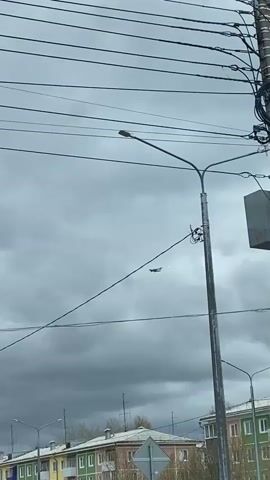 The video shows a cloudy sky above a street in what looks like a residential area. Power lines stretch across the top of the frame. In the middle ground, a street light stands tall, and below it, a multi-colored apartment building lines the street. A road sign is visible on the lower right. A dark shape, apparently an airplane, is seen moving through the clouds, getting further away from the camera. The overall scene presents a dull, overcast day in an urban environment.