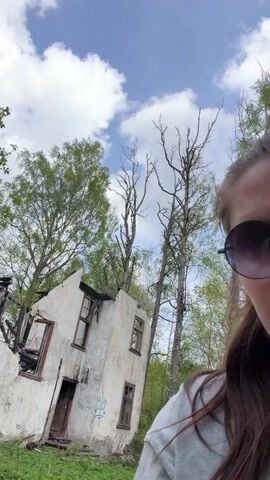 The video shows a woman wearing sunglasses, partially visible on the right side of the frame, with a ruined building in the background. The building is a two-story structure with a white exterior, heavily damaged by fire. The roof is partially collapsed, and the windows are broken or missing. The charred remains of the building's interior are visible. Trees surround the building, some with green leaves and others with bare branches. The sky is blue with scattered white clouds. The camera appears to be handheld, moving slightly as the person filming walks or adjusts their position.