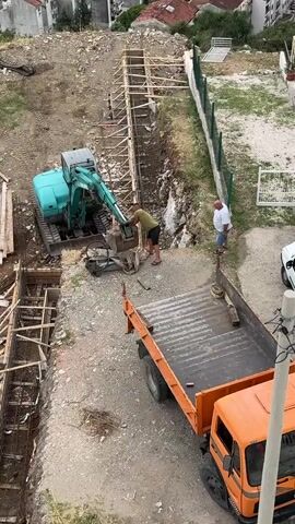This aerial view captures a construction scene in Sušćepan, Montenegro, likely during daylight hours given the shadows. Two men are actively involved in the groundwork. One man, wearing a green shirt and dark shorts, operates a small, turquoise excavator, appearing to scoop material from a trench. He is also seen holding a shovel. The second man, in a white shirt and blue shorts, stands near the excavator, observing the work.

The setting is a sloped construction site. A deep trench, reinforced with wooden forms and rebar, runs horizontally across the lower portion of the frame. The excavator is positioned at one end of this trench, digging into the earth. Further up the slope, a similar wooden framework suggests another section of the foundation or retaining wall is being prepared. To the right of the trench, an orange flatbed truck is parked, its bed empty. A white car is visible further up the hill. The surrounding area shows signs of residential development, with buildings visible in the background. The overall atmosphere is one of active construction and labor, with no discernible emotional expressions from the individuals. There is no visible text in the media file.