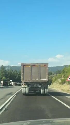 A rear view of a dump truck driving on a paved road. The truck is brown and has three large compartments in the back. It has dual rear wheels on each side. The road has white lane markings. On the right side of the road, there are trees and a steep embankment. There is a triangular road sign with a black symbol on it, indicating a curve. On the left side of the road, there are more trees and some structures that appear to be buildings or stalls. The sky is blue with some scattered clouds. The overall scene is that of a sunny day on a rural highway.