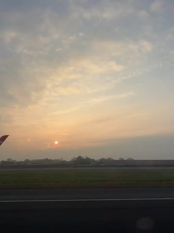 The video shows a view from inside an airplane, looking out over a runway at sunrise. The sun is visible as a bright circle in the sky, partially obscured by clouds. A small part of the airplane's wing is visible in the lower left corner. Beyond the runway is a green field with trees in the distance, partially shrouded in fog. The sky is filled with a mix of white and gray clouds.