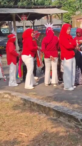 A group of individuals dressed in red tops and white pants, some wearing red headscarves and white feathered headdresses, are gathered outdoors. They appear to be participating in a parade or performance, with some holding drums or other musical instruments. A younger child in a white uniform and patterned skirt is also present. In the foreground, two young boys are visible; one is in a camouflage military uniform with a green beret, and the other is wearing a pilot's hat and uniform. The background shows a building with a corrugated metal roof and a tree. The ground is covered in dry grass and dirt. The overall scene suggests a festive or ceremonial event taking place during the daytime.