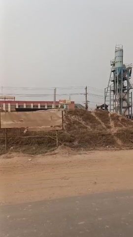 The video shows a static shot of a dusty, rural scene. In the background, there's a large, multi-story industrial structure, possibly a concrete mixing plant, with a tall silo on top. There are various pipes, stairs, and other industrial components visible on the structure. To the left of the structure is a long, low building with a red roof.

The foreground is a dirt road. A man on a bicycle rides along the road from right to left, away from the camera. Two women wearing traditional clothing walk on the same dirt road, from right to left as well.

In the area between the road and the industrial structure, there are numerous pieces of metal scrap, piles of dirt, and other debris. The sky is overcast and hazy, suggesting a dull, overcast day.
