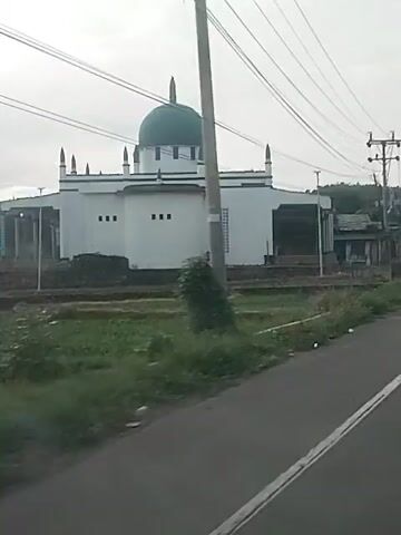 The video shows a mosque with a green dome and minarets, seen from a moving vehicle. The mosque is white with some dark accents. There are power lines and utility poles in the foreground and background, and some greenery. The video appears to be taken during the daytime, under an overcast sky. The vehicle moves past the mosque, showing its entrance gate which has Arabic writing.