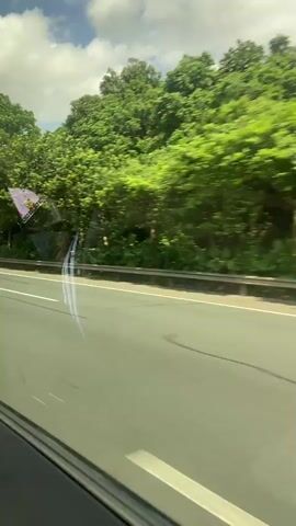 The video shows a view from inside a vehicle moving along a highway. The highway has multiple lanes and a white barrier separating the lanes from the roadside. The roadside is lined with dense green trees and vegetation. In the sky, there are white clouds against a blue background. A green sign with "50" is visible on the roadside, indicating a speed limit. There are no people or animals visible in the video. The camera appears to be static relative to the vehicle's interior.