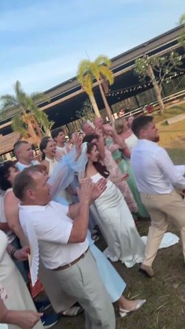 The video captures a lively outdoor celebration, likely a wedding reception, under a bright, clear sky. In the center of the frame, a man, presumably the groom, wearing a white long-sleeved shirt and light beige trousers, is enthusiastically spraying champagne from a bottle. He is in a dynamic, slightly crouching posture, directing the spray towards the right side of the frame.

To his immediate left, a woman, likely the bride, is visible in a white wedding gown, with a distinctive light blue sheer fabric or veil around her head and shoulders. She is smiling broadly and clapping her hands, looking towards the groom.

Surrounding the couple is a group of joyful guests, all clapping, cheering, and smiling. Their expressions convey excitement and happiness. One man in a white polo shirt and beige trousers is prominently in the foreground on the left, clapping with gusto. The guests are dressed in various semi-formal attires, including dresses in different colors and light-colored shirts.

The event takes place on a well-maintained grassy area. In the background, several tall palm trees are visible, suggesting a warm climate or tropical setting. Further back, a large building with a dark roof and decorative string lights can be seen, possibly an event hall or reception area. To the right of the groom, on the grass, there is a small hay bale draped with an orange cloth. The overall atmosphere is festive and full of celebratory energy.