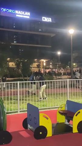 The video shows a park or playground scene at night in Kiryat Motzkin, Israel. In the foreground, there is red-surfaced playground equipment, including yellow and blue train-like structures and a green tunnel or barrel. A white fence separates this area from a grassy patch and what appears to be a running track.

Beyond the fence, several people are visible walking and standing around. The area is illuminated by streetlights. In the background, modern buildings rise, with one prominently displaying the illuminated sign "FORID NADLAN" and another sign with Hebrew text that reads "שפע" (Shefa). The sky is dark, indicating it is nighttime. No specific activities are ongoing with the playground equipment, but people are casually moving about in the background. The overall atmosphere appears calm.