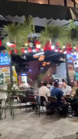 The video depicts an outdoor seating area of a restaurant or bar at night. The area is decorated with hanging plants, string lights, and small red flags. Several groups of people are seated at tables, engaging in conversation and eating. The area is covered by a partial roof, and there are buildings visible in the background. The floor is paved with interlocking stones.