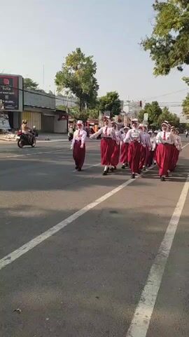 A group of female students in uniforms are marching in formation on a paved road. They are wearing white shirts, red skirts, and white hats. Some of them are holding small flags. The students are walking from left to right in the frame. In the background, there are buildings, trees, and a motorcycle. The sky is clear and the sun is shining. There are white lines painted on the road. The students appear to be participating in a parade or a similar event. One student in the middle of the group has the number 012 visible on her shirt.