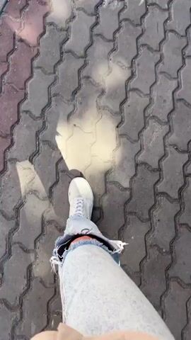 The video shows a first-person perspective of someone walking outdoors in Tolyatti, Russia. The person is wearing light-wash distressed jeans and white sneakers. The ground is a mix of patterned brick pavers and asphalt, suggesting a sidewalk or pedestrian area. Sunlight casts shadows, indicating it is daytime, likely sunny. Some sparse vegetation is visible along the edge of the path. The overall impression is a casual walk on a warm day.