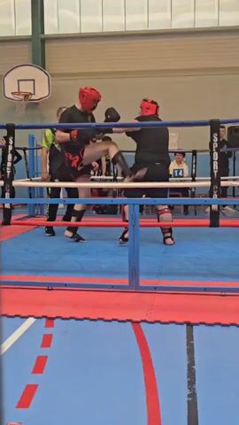 Two boxers wearing headgear are in a boxing ring. A referee separates them and picks up a fallen boxing glove. A basketball hoop is visible in the background. There are spectators outside the ring. The floor outside the ring is marked with red lines.