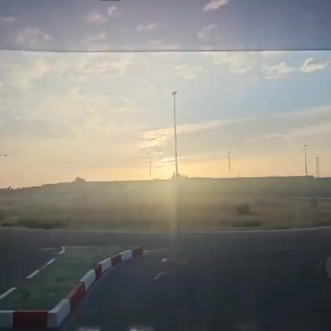 The video begins with a view of a street lamp and a sunset over a field. In the distance, there are power lines and a structure that resembles an industrial building. There are also some figures that appear to be animals grazing in the field. The foreground shows a portion of a road with red and white markings, suggesting a traffic island or a road divider. The sun is low in the sky, casting a warm glow and creating a lens flare effect. The sky is filled with clouds. As the video progresses, the view shifts slightly, but the overall scene remains consistent. The figures in the field become slightly more discernible, appearing to be a few animals, possibly sheep or goats, scattered across the grassy area. The light from the setting sun continues to illuminate the landscape. The industrial structure in the distance remains visible, silhouetted against the sky. The street lamp in the foreground continues to be a prominent feature.