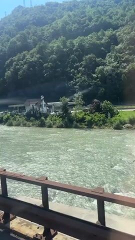 The video captures a scenic view from what appears to be a vehicle, possibly a train or car, moving alongside a wide, greenish river. The river is fast-flowing with visible turbulence and foam. On the opposite bank, there is a lush, green mountainside covered in dense trees. A white building, possibly a house with a distinct white steeple or pointed roof, is situated near the riverbank, partially obscured by trees. A road or pathway runs behind the building, with some structures that might be covered parking or a small commercial area. In the distance, on the upper slopes of the mountain, tall electricity pylons are visible against a bright blue sky with sunlight casting shadows. The foreground shows a railing or barrier, likely from the vehicle's vantage point, which is dark brown and appears to be made of metal. The video's camera movement suggests the vehicle is traveling, indicated by the blurred foliage and the shifting perspective of the landscape. The overall impression is of a journey through a natural, mountainous region with a river as a prominent feature.