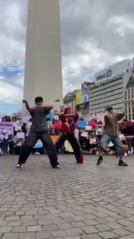 The video shows three dancers performing in front of a crowd of seated people holding signs in a plaza in Buenos Aires, Argentina. The tall Obelisco de Buenos Aires is visible behind them, along with a SIGLO shopping center. The dancers are wearing street clothes; one is wearing red and black, and the other two are wearing grey, black and tan clothing. As the first dancer walks toward the seated group, a large bird flies over the audience holding signs. The crowd is a mixture of men and women, seated on the cobblestones of the plaza. They are holding signs that contain photos and writing. The weather looks overcast.