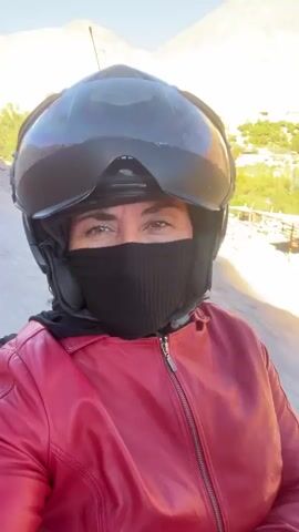 A woman wearing a red leather jacket, a black face mask, and a black helmet with a clear visor is looking directly at the camera. She appears to be on a moving vehicle, possibly a motorcycle or scooter, as indicated by the slight motion blur and the background of a dry, mountainous landscape. The sun is shining brightly, casting a shadow of the woman and her vehicle on the ground. The environment appears to be arid and rural, with some sparse vegetation and buildings visible in the distance.