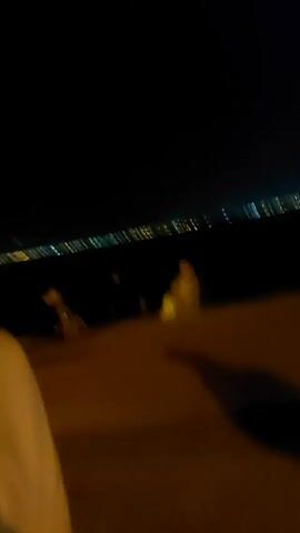 Here is a concise description of the video:

The video begins with a close-up of a man's face, wearing glasses, with a blurred city skyline visible in the background. The camera then moves to show a group of people walking along a paved area at night. The people are mostly seen from the back. The lighting is dim and appears to be from streetlights and distant city lights.