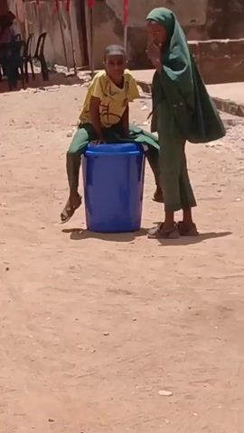 The video shows two children in an outdoor setting. A young boy is seated on a blue plastic bucket, wearing a yellow shirt with green pants and sandals. A young girl, clad in a green hijab and matching green outfit with sandals, is standing beside him. The background includes buildings, a red banner, and a scattering of litter on the ground. A man on a moped then passes behind the boy. The environment appears to be a residential or community area with a sandy or dirt ground.