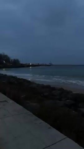 The video shows a coastal scene on a cloudy, overcast day, likely at dusk or dawn. A calm sea with small waves laps against a sandy beach protected by a rocky barrier. In the distance, a structure or pier with lights can be seen extending into the water. A concrete walkway runs along the edge of the scene in the foreground. There are no people visible in the video.