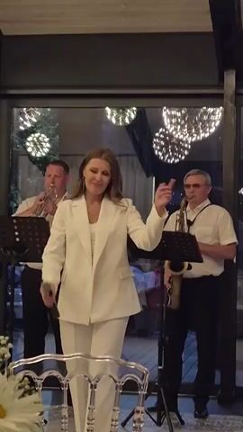 A woman in a white pantsuit is singing into a microphone, holding it with her right hand. She has long, light brown hair and is wearing a white top under her jacket. Her left hand is slightly raised, with her fingers extended. Behind her, to her left, a man in a white shirt is playing a trumpet. To her right, another man in a white shirt and a yarmulke is playing a saxophone. Both men have music stands in front of them. The scene appears to be indoors, possibly a restaurant or event venue, with a large glass window or door in the background. There are decorative lights hanging from the ceiling. In the foreground, a portion of a table with white flowers and clear chairs is visible. The overall atmosphere suggests a live musical performance.