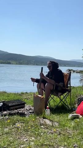 A man is sitting in a camping chair by the water on a bright, sunny day. He wears a black hoodie, sunglasses, and shorts, and appears to be whittling a stick with a tool. Beside him on the grassy ground are a brown paper bag, a clear plastic bottle, and a red bag. A small portable grill is visible to his left. The scene shows a wide body of water, possibly a river or lake, with distant green hills under a clear blue sky. The ground is covered in green grass with patches of dandelions. The activity takes place in Слизнево, Russia.