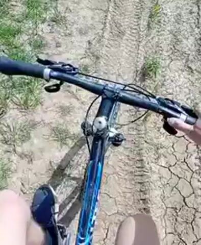 The video shows a first-person view of someone riding a blue mountain bike on a muddy, dirt road next to a field of green grass. The road has tire tracks and some patches of water. In the distance, there are rolling hills and trees under a clear, blue sky. The person riding the bike is wearing a short-sleeved shirt and has their leg visible in the bottom right of the frame.