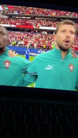 The media file is a video recording of a soccer match, viewed on a laptop screen. The subjects are several male soccer players wearing teal track jackets with the Portuguese national team crest. They appear to be singing or chanting, with open mouths and expressive faces, indicating strong emotions like pride or passion. The players are standing on a soccer field, with their arms around each other's shoulders. In the background, a large stadium filled with spectators wearing red is visible. The stands are packed, suggesting a significant event. The sky appears to be bright, implying it's daytime, though the exact time of day is not discernible. There is visible text on a banner in the background that reads "FIFA WORLD CUP 2026". The overall scene depicts a moment of camaraderie and national pride during a major sporting event. The presence of the laptop screen indicates the video is being watched, possibly in a casual setting at home or on the go. The provided location context of Nikopol, Bulgaria, is not directly supported by any visual cues within the video itself, as the setting is clearly a large international stadium.