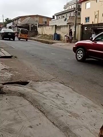 The video shows a residential street. Initially, a maroon SUV drives down the street. Then, a silver sedan follows. Finally, a black sedan with chrome rims passes. On the right side of the street, a person stands near a table with goods displayed. Two people are seated in chairs nearby. The buildings are low-rise and appear to be located in a residential area. The day is overcast. There is a deep pothole in the pavement near the bottom of the video.