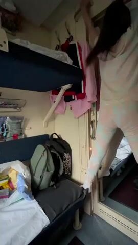 A person wearing pajamas with a tie-dye pattern of pink and white is climbing into a bunk bed on a train. The bunk bed is dark blue and has white bedding. The person's legs are extended as they grasp a metal bar to pull themselves up. Several items of clothing, including a pink sweatshirt and a red and black jacket, are hanging on hooks near the bunk bed. Luggage, including a green backpack and a dark gray bag, is visible below the lower bunk. The train compartment appears to be narrow, with wood-paneled walls and additional bunk beds visible in the background.