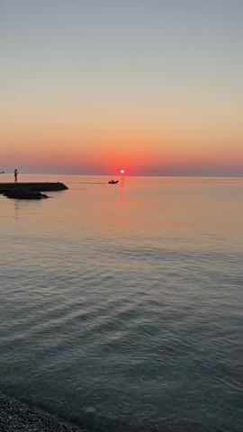 A wide shot captures a serene ocean scene at sunset. The sun, a vibrant red orb, hangs low on the horizon, casting a warm glow across the water and sky. The water is calm, with gentle ripples reflecting the colors of the sunset. A small inflatable boat with a single person on board is moving across the water from left to right, leaving a slight wake. In the distance, on the left side of the frame, a person stands on a rocky pier, silhouetted against the fading light. The sky transitions from a soft pink and orange near the horizon to a pale blue overhead. The overall atmosphere is peaceful and tranquil.